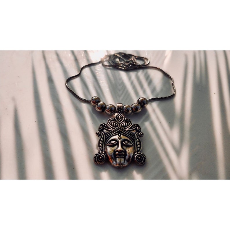 May include: A silver necklace with a detailed pendant featuring a face. The pendant is ornate and has a crown-like design. The necklace is on a white surface.