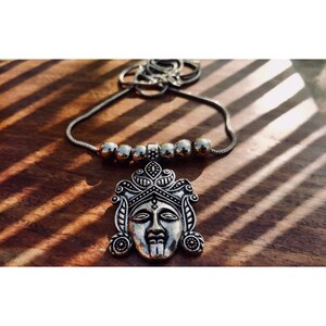 May include: A silver necklace with a detailed face pendant and silver beads. The necklace is on a wooden surface.