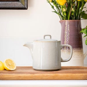 Pu&ograve; includere: Una teiera in ceramica beige chiaro con coperchio e manico, su un tagliere di legno. Dietro c'&egrave; un brocca viola e bianca con fiori gialli. C'&egrave; anche mezzo limone sul tagliere.