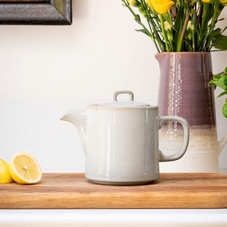 Pu&ograve; includere: Una teiera in ceramica beige chiaro con coperchio e manico, su un tagliere di legno. Dietro c'&egrave; un brocca viola e bianca con fiori gialli. C'&egrave; anche mezzo limone sul tagliere.