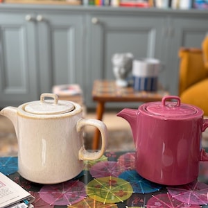 May include: Two ceramic teapots with lids and side handles. One teapot is a speckled off-white colour with a smooth finish, while the other teapot is a solid, glossy deep magenta colour. Both feature a cylindrical body and a short, angled spout.