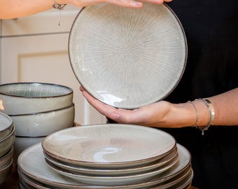 May include: A set of round ceramic plates and bowls featuring a light grey glaze with a subtle, radiating sunburst pattern of thin, etched lines and a dark, contrasting rim on each piece, stacked neatly on a wooden surface.