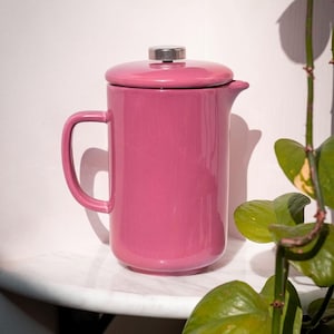 Puede incluir: Una cafetera de prensa francesa rosa con tapa de acero inoxidable. La cafetera es cilíndrica con asa y boquilla. El fondo es blanco con una planta en la esquina.