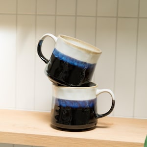 Könnte beinhalten: Zwei gestapelte Keramikbecher mit zweifarbigem Design, bestehend aus einer glänzend schwarzen Basis und einem cremeweißen oberen Bereich. Ein leuchtend blaues Glasurband trennt die beiden Farben, jeder Becher hat einen geschwungenen Henkel.