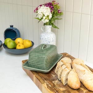 May include: A rectangular sage green ceramic butter dish with a ribbed lid and matching base, positioned on a wooden cutting board next to a sliced baguette. In the background, a blue bowl holds limes and lemons, and a white speckled vase holds flowers.