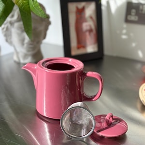 May include: A solid pink ceramic teapot with a matching lid and a removable stainless steel mesh tea infuser basket. The teapot features a cylindrical body, a short spout, and a curved handle, presented with the lid placed beside the infuser.