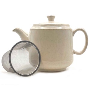 Pode incluir: Uma chaleira de cerâmica bege com um infusor de chá de aço inoxidável. A chaleira tem uma tampa e uma alça. O infusor é separado e tem um design de malha.
