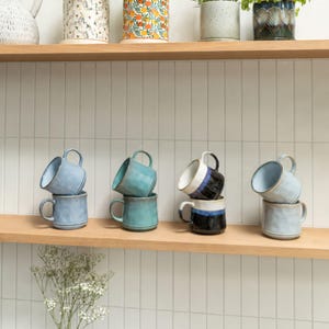 May include: Eight ceramic mugs arranged in four stacks of two on a wooden shelf. The mugs feature various glaze finishes including light blue, teal, and a two-tone black and white design. Each mug has a rounded handle and a textured, slightly irregular surface.