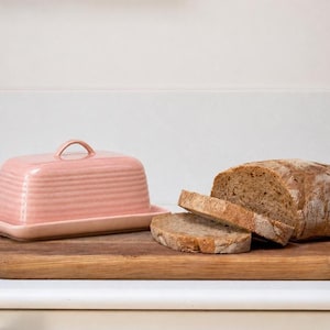 Puede incluir: Una mantequillera rectangular de cerámica rosa con tapa a juego que presenta un pequeño asa arqueada y detalles acanalados horizontales, situada junto a una hogaza de pan integral con dos rebanadas cortadas sobre una tabla de madera.