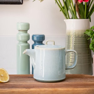 May include: A light blue ceramic teapot with a lid and handle sits on a wooden surface. Two decorative ceramic bottles in shades of blue and green are behind the teapot. A vase with flowers is in the background.