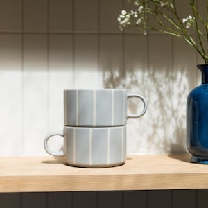 May include: Two stacked light grey ceramic mugs featuring vertical white stripes. Each mug has a rounded handle on the side and a cylindrical body. The mugs are positioned together, creating a uniform appearance with consistent spacing between the stripes.