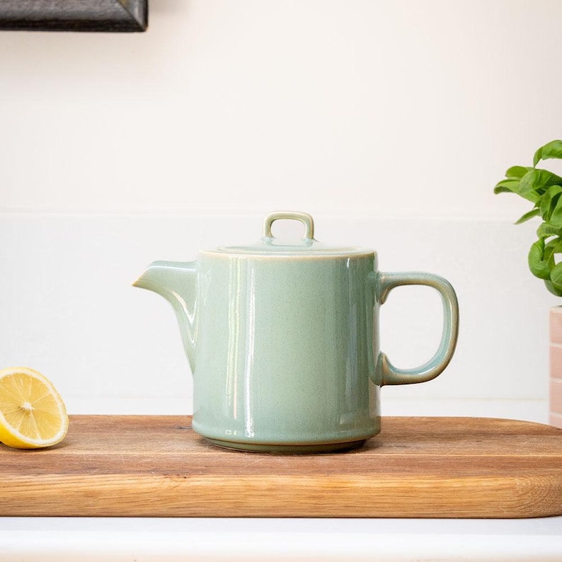 Pu&ograve; includere: Una teiera in ceramica verde chiaro con coperchio e manico. La teiera &egrave; su un tagliere di legno con met&agrave; limone. La teiera ha un beccuccio per versare il t&egrave;.