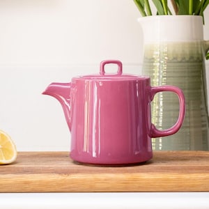 Può includere: Una teiera in ceramica lucida color magenta, con coperchio e manico. La teiera è appoggiata su un tagliere di legno. Una metà di limone è visibile a sinistra. Un vaso bianco e verde è sullo sfondo.
