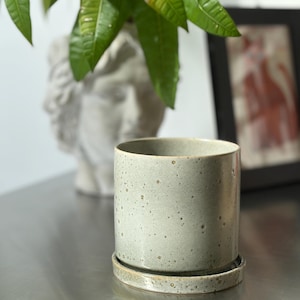 May include: A cylindrical ceramic planter with a speckled light grey finish and a matching circular saucer base. The pot features a smooth, straight-sided design with subtle variations in the glaze texture, creating a neutral and functional vessel.