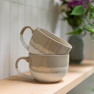 Op de afbeelding: Twee gestapelde keramische mokken met een tweekleurig ontwerp, bestaande uit een glad beige bovendeel en een gespikkeld wit onderdeel. De mokken hebben een ronde vorm, een brede opening en een gebogen handvat aan de zijkant, met een duidelijke horizontale overgangslijn tussen de twee kleuren.