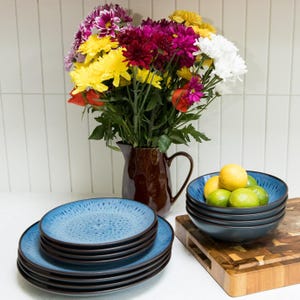 May include: A set of blue and black speckled ceramic plates stacked in a pile next to a stack of matching blue and black bowls filled with yellow lemons and green limes, with a dark brown ceramic pitcher holding a bouquet of colourful flowers.
