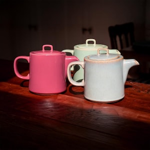 May include: Three ceramic teapots in pink, green, and blue-grey sit on a wooden surface. Each teapot has a handle, a spout, and a lid with a small handle. The teapots are arranged in a row, with the pink one in the foreground.