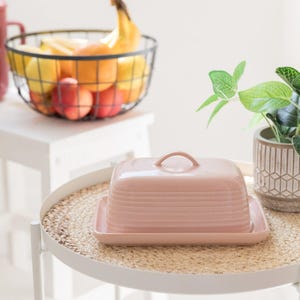 May include: A rectangular, light pink ceramic butter dish with a matching lid featuring a curved handle. The dish has horizontal ribbed detailing along the sides and sits on a flat, rectangular base, presenting a smooth, glossy finish throughout.