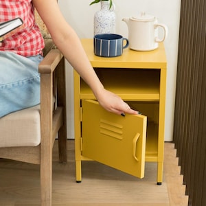 Puede incluir: Una mesita de noche de metal color amarillo mostaza con un estante superior abierto y un armario inferior con puerta abatible. La puerta incluye tres ranuras de ventilación horizontales y un tirador vertical. Una taza azul a rayas descansa encima.