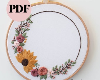 May include: A beginner embroidery hoop with a floral wreath design. The wreath features a sunflower, pink roses, and green leaves. The hoop is made of wood and has a white fabric background. The text "Beginner" is printed below the hoop.