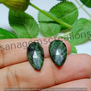 Może przedstawiać: Dwa fasetowane, łezkowate kamienie szlachetne. Kamienie są w głębokim odcieniu zieleni z ciemniejszymi wtrąceniami i mają wypolerowaną, odbijającą światło powierzchnię. Kamienie są prezentowane na tle zielonych liści i dłoni.