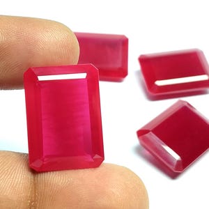 May include: Four rectangular, faceted, vibrant pinkish-red gemstones. Each stone features a clear, polished surface with sharp, beveled edges that catch the light, creating bright white reflections on the top facets of the translucent, deep red material.