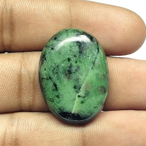 Könnte beinhalten: Oval geformter grüner Edelstein mit schwarzen und braunen Einschlüssen. Der polierte Stein hat eine glänzende Oberfläche und wird in einer Hand gehalten. Der Edelstein wird wahrscheinlich für Schmuck oder andere dekorative Zwecke verwendet.