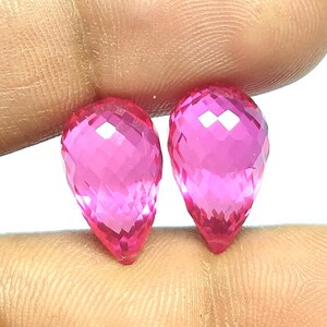 May include: Two pear-shaped, faceted gemstones with a vibrant, translucent hot pink color. Each stone features a pointed bottom and a rounded top, with a geometric cut that reflects light across the surface of the bright pink, teardrop-shaped gems.