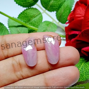May include: Two oval-shaped, pink gemstones with faceted tops. The gems are held in a hand, with a red rose and green leaves in the background. The text "sanagemscreations" is visible on the image.