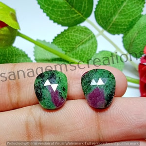Op de afbeelding: Twee edelsteen cabochons in groen, rood en wit. De edelstenen hebben een unieke vorm en een facetontwerp. De afbeelding is geplaatst tegen een achtergrond van groene bladeren en een rode roos.