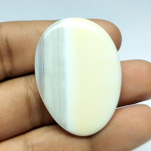 Könnte beinhalten: Oval geformter Edelstein mit polierter Oberfläche. Der Stein hat eine cremeweiße Farbe mit einem vertikalen Band aus blassen Grau- und Lavendeltönen. Der Edelstein wird in einer Hand gehalten, um seine Größe und Form zu zeigen.