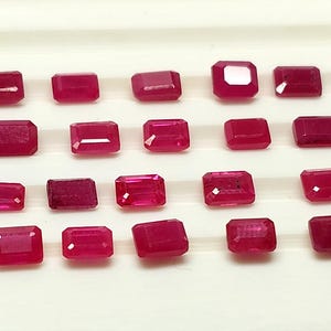 May include: A collection of twenty loose, rectangular-cut rubies arranged in four rows of five. Each gemstone displays a deep, vibrant red hue with faceted surfaces that reflect light, showcasing a consistent shape and polished finish throughout.
