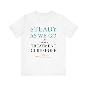 Puede incluir: Camiseta blanca con el texto "STEADY AS WE GO" en color turquesa, seguido de "TREATMENT CURE HOPE" en negro. Un pequeño gráfico de rosa está entre las frases. El logotipo de Team Fox está en la parte inferior.