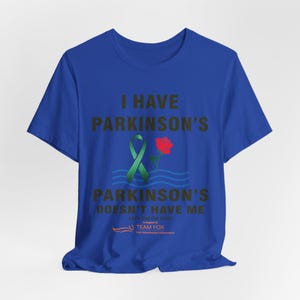 Puede incluir: Camiseta azul real con el texto "I Have Parkinson's, Parkinson's Doesn't Have Me". Un lazo verde y una rosa roja aparecen en el diseño. La camiseta también incluye el logotipo de Team Fox.