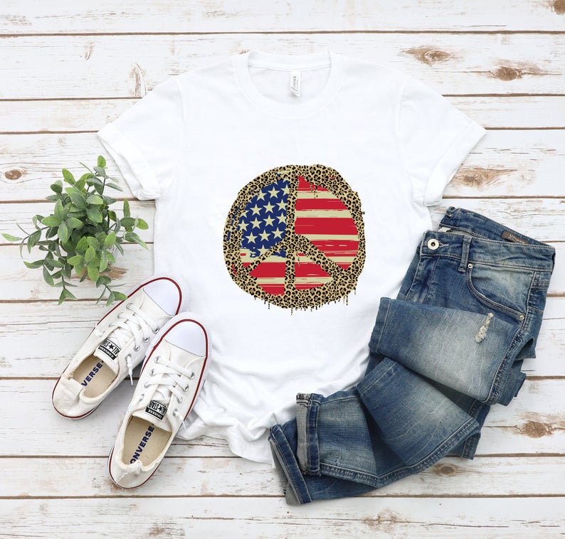 May include: White t-shirt with a peace sign design. The peace sign is made up of an American flag with a leopard print overlay. The shirt is laid out on a woodgrain surface with a pair of white sneakers and blue jeans.