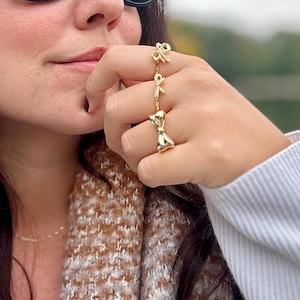 Può includere: Una mano di donna con tre anelli d'oro con motivi a fiocco. Gli anelli sono impilati sul suo dito. Gli anelli sono un accessorio di moda popolare.