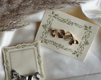 Op de afbeelding: Twee paar gouden en zilveren oorringen in de vorm van een strik, gepresenteerd op een witte kaart met een decoratieve rand.