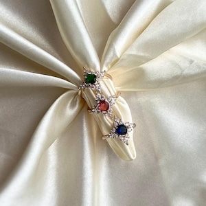 Op de afbeelding: Drie gouden ringen met een stervormig ontwerp. Elke ring heeft een edelsteen in een andere kleur in het midden: groen, rood en blauw.