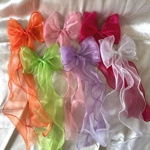 Könnte beinhalten: Eine Sammlung farbenfroher Organza-Haarschleifen in Orange, Rosa, Grün, Lila und Weiß. Jede Schleife hat lange, fließende Bänder, die einen skurrilen und eleganten Look erzeugen. Die Schleifen sind auf einem weichen, weißen Stoff angeordnet.