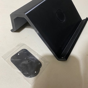 May include: Black plastic stand with a raised platform and a circular cutout. The stand is designed to hold a tablet or phone upright. A small black plastic piece is included in a clear plastic bag.