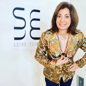 Op de afbeelding: Een vrouw draagt een gouden blazer met een bloemenpatroon en daaronder een roze T-shirt. De blazer heeft een knoopsluiting en een zak aan de linkerkant. De vrouw staat voor een witte muur met een zwart logo waarop "Susan Elia" staat.