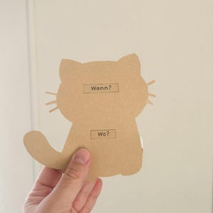 Pode incluir: Um recorte de cart&atilde;o castanho de um gato com as palavras "Wann?" e "Wo?" escritas nele.