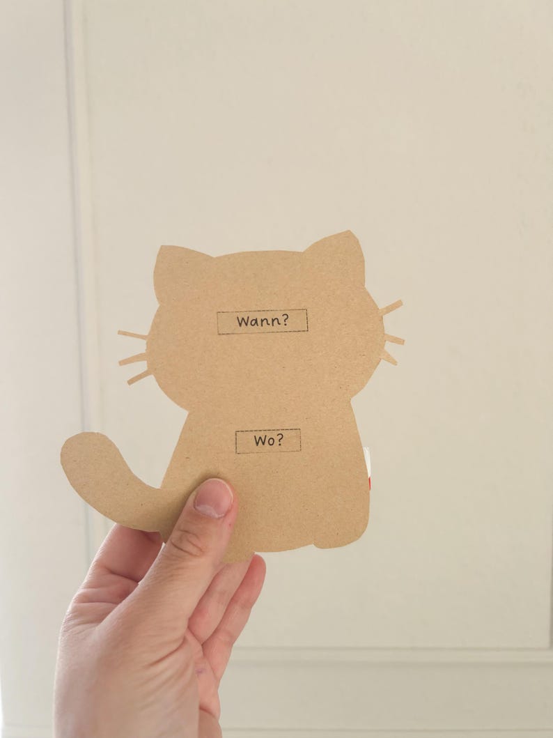 Pode incluir: Um recorte de cart&atilde;o castanho de um gato com as palavras "Wann?" e "Wo?" escritas nele.