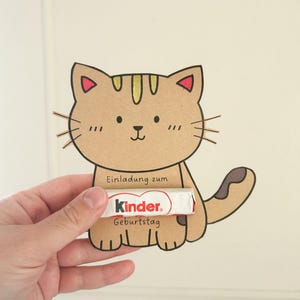 Pode incluir: Um gato de desenho animado castanho com listras amarelas e orelhas vermelhas segura uma barra de chocolate Kinder que diz "Einladung zum Geburtstag" (Convite de anivers&aacute;rio).