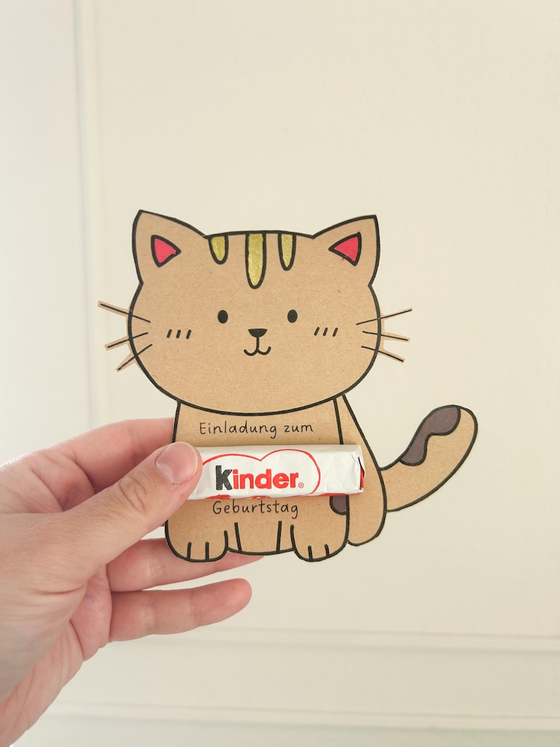 Pode incluir: Um gato de desenho animado castanho com listras amarelas e orelhas vermelhas segura uma barra de chocolate Kinder que diz "Einladung zum Geburtstag" (Convite de anivers&aacute;rio).