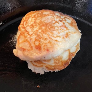 Może przedstawiać: Zbliżenie dwóch złocistobrązowych naleśników, ułożonych jeden na drugim. Naleśniki mają lekko nierówną powierzchnię i lekką, puszystą konsystencję. Leżą na ciemnej powierzchni, prawdopodobnie żeliwnej.