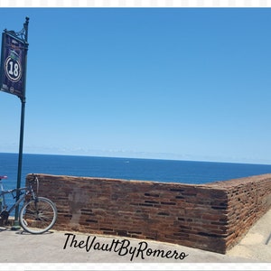 May include: A brick wall with a blue sky and ocean in the background. A bicycle is leaning against the wall. A small tower with a white dome is on the right side of the image. Two people are standing on the wall, one is taking a picture of the other. The sign on the pole reads "Route 18".