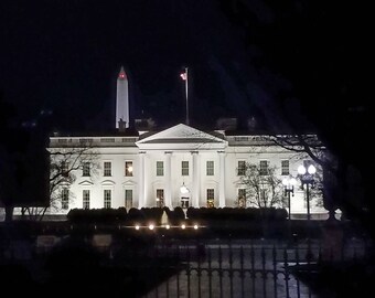 May include: A nighttime view of the White House, illuminated by bright lights. The building is white with a black roof and many windows. The Washington Monument is visible in the background.