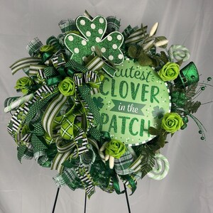 May include: A St. Patrick's Day wreath with green ribbons, shamrocks, and roses. The wreath has a sign that reads "Cutest Clover in the Patch". The wreath is displayed on a black stand.