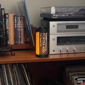 May include: A Kodak Professional PORTRA 400 color negative film canister stands on a wooden shelf, next to books and audio equipment. The canister is black with orange and white text. A record player and stereo components are in the background.